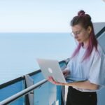 A young woman works remotely on her laptop by the sea on a sunny day, embodying the digital nomad lifestyle.